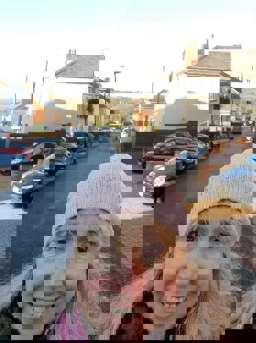 woman with long blonde hair wearing pink woolly hat with residential street behind her